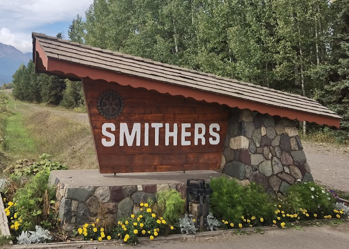 Wooden Smithers sign with a shingled roof and Rotary emblem on a stone base, with yellow flowers in front and aspen trees behind.